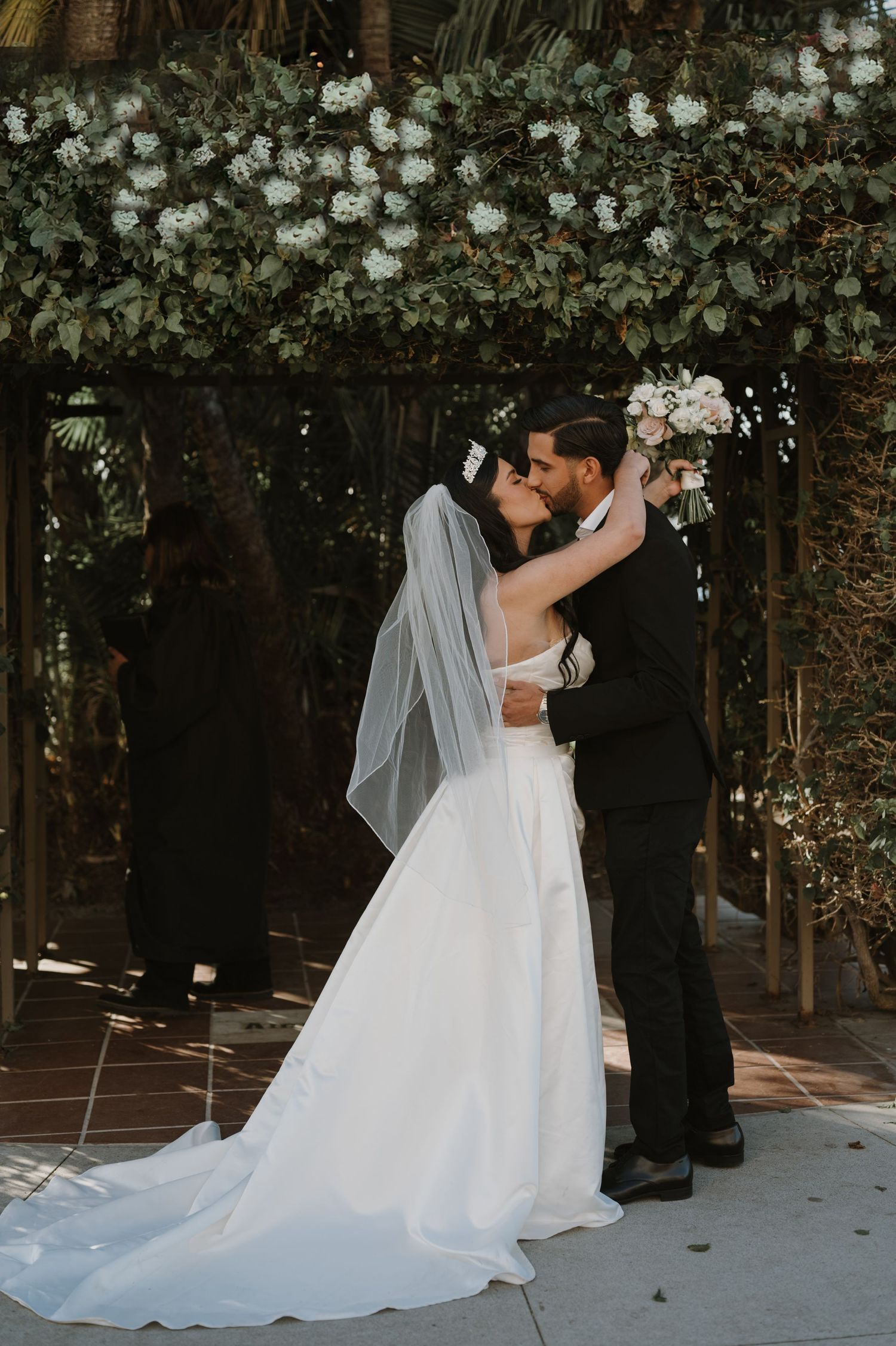 Newlyweds share their first kiss under an arch decorated with white flowers.