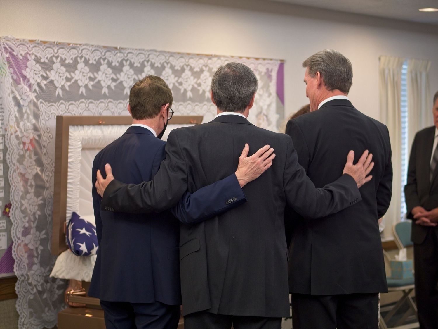 Three men stand in front of the casket of their aged father with their arms around each other