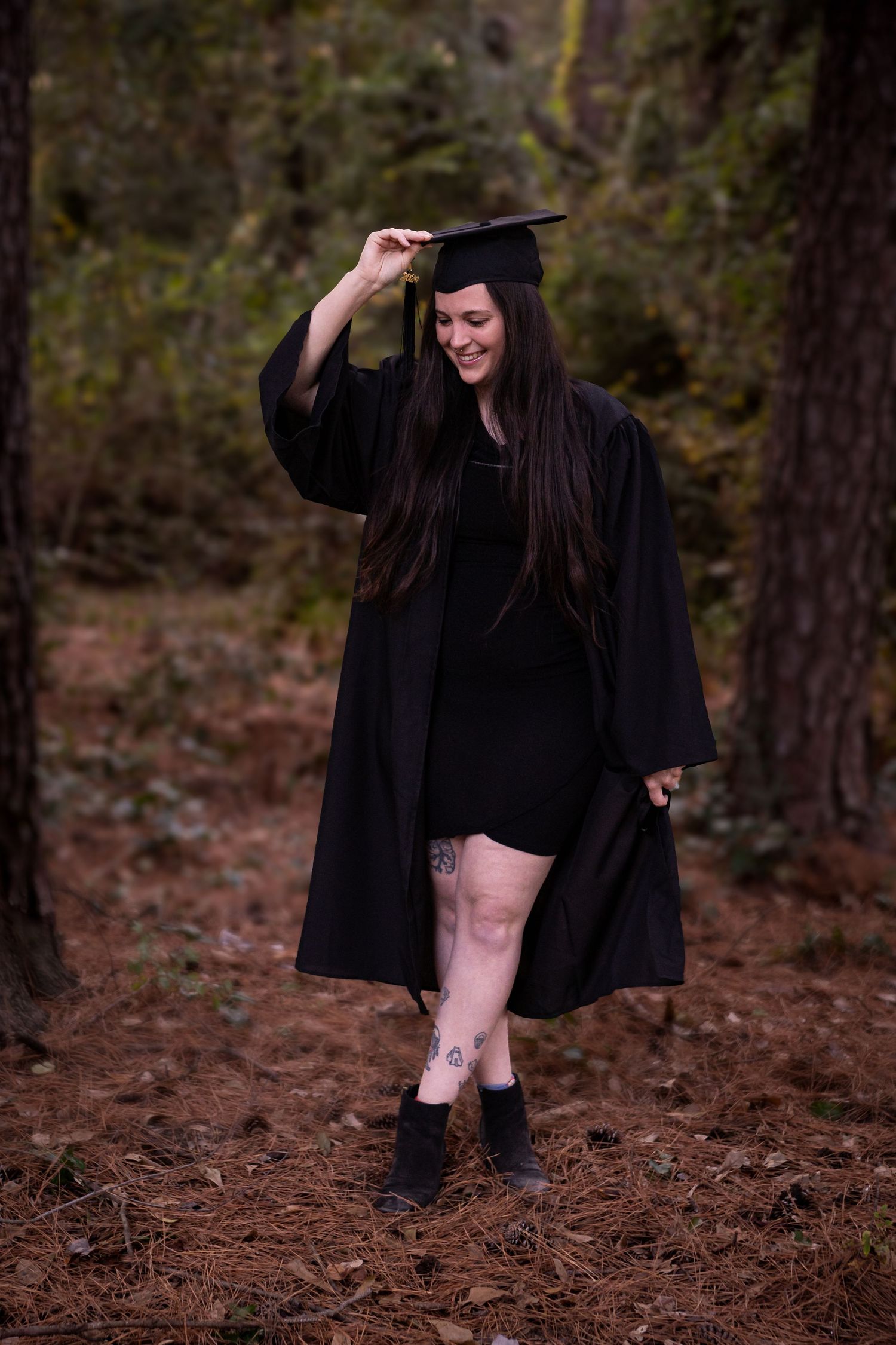 Graduate in black cap and gown poses playfully in a forest setting.