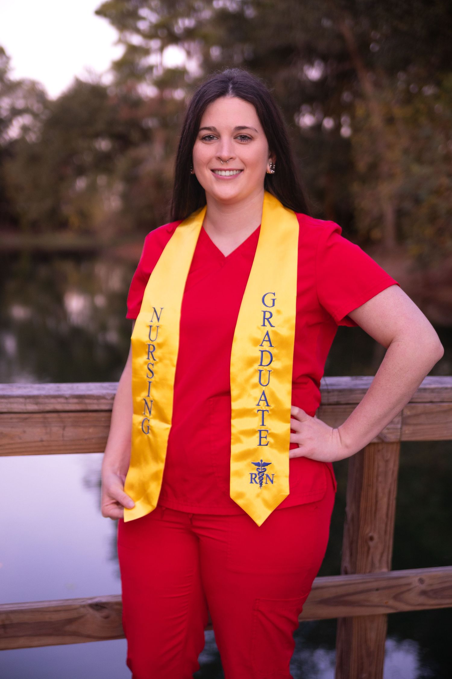 Graduate wearing red outfit and yellow stole poses on wooden deck.
