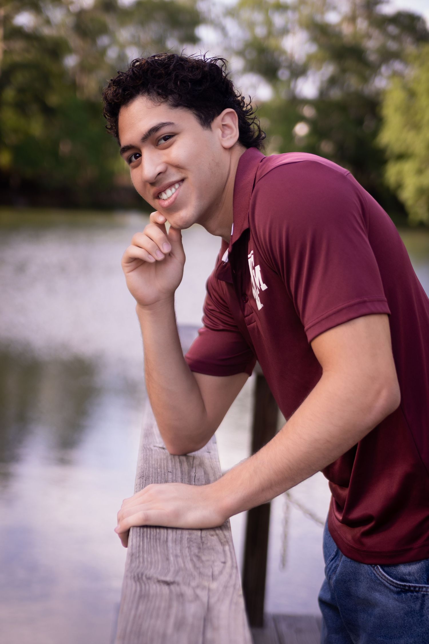 A Senior in a burgundy t-shirt leans thoughtfully against a wooden post near a serene lake in Houston Texas at Kickerillo Misher Preserve