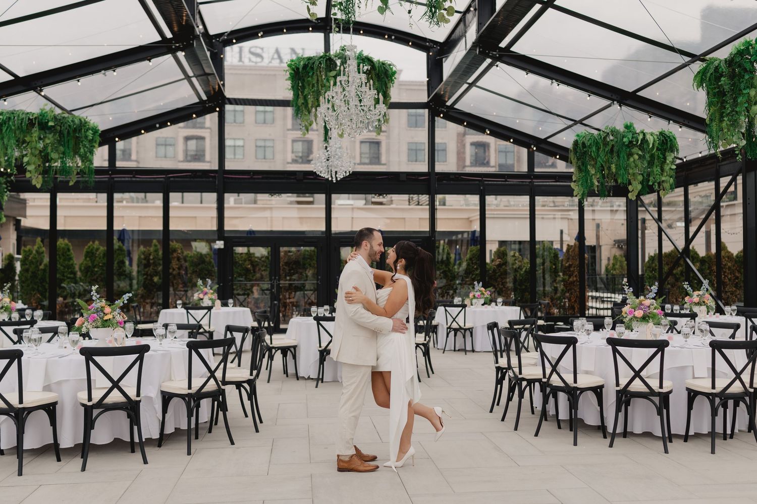 Couple embraces and dances in elegant glass greenhouse wedding venue with chandeliers and white-draped reception tables.