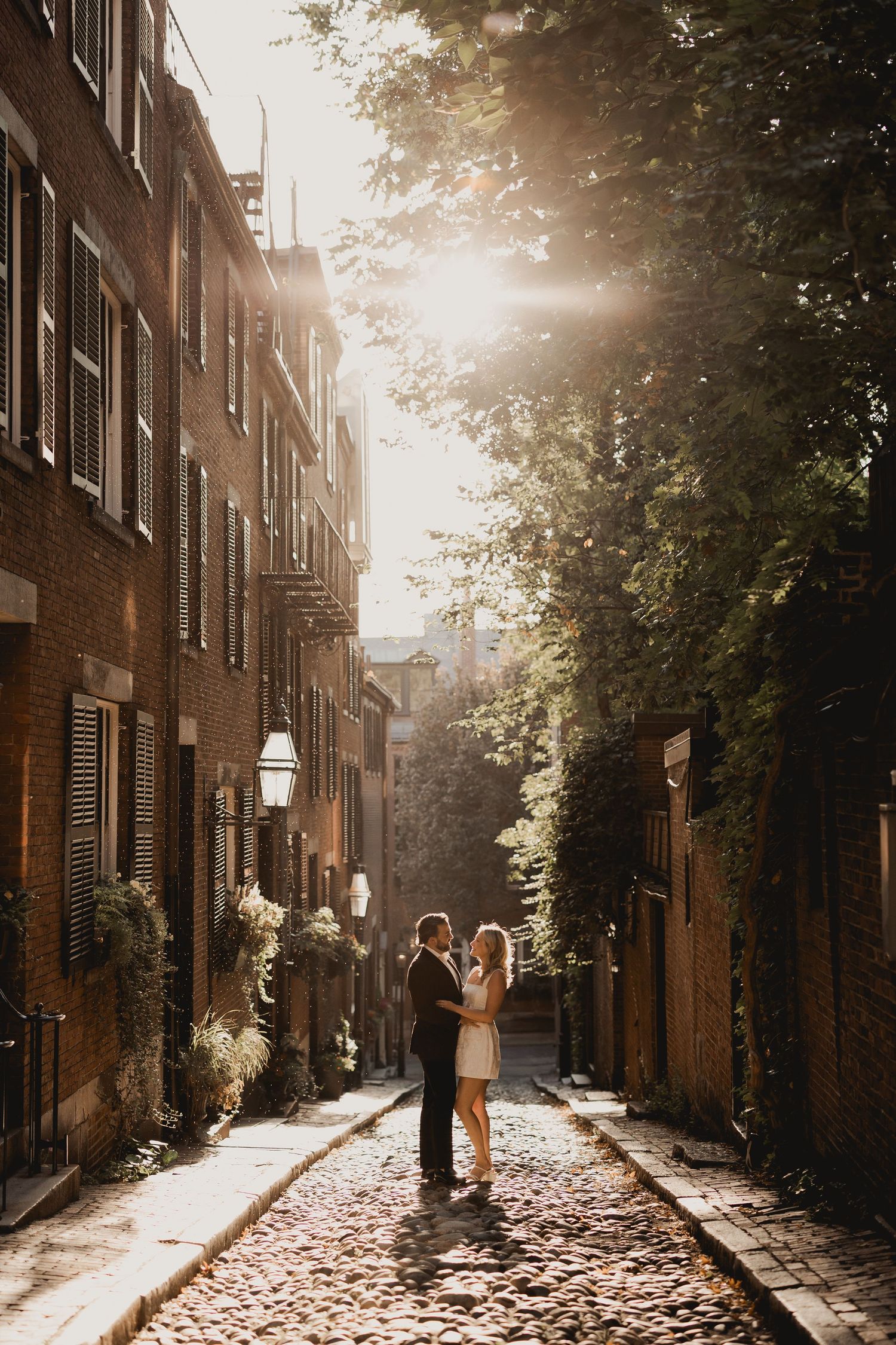 Couple embraces on sunlit cobblestone alley in Boston's Beacon Hill during golden hour engagement session.