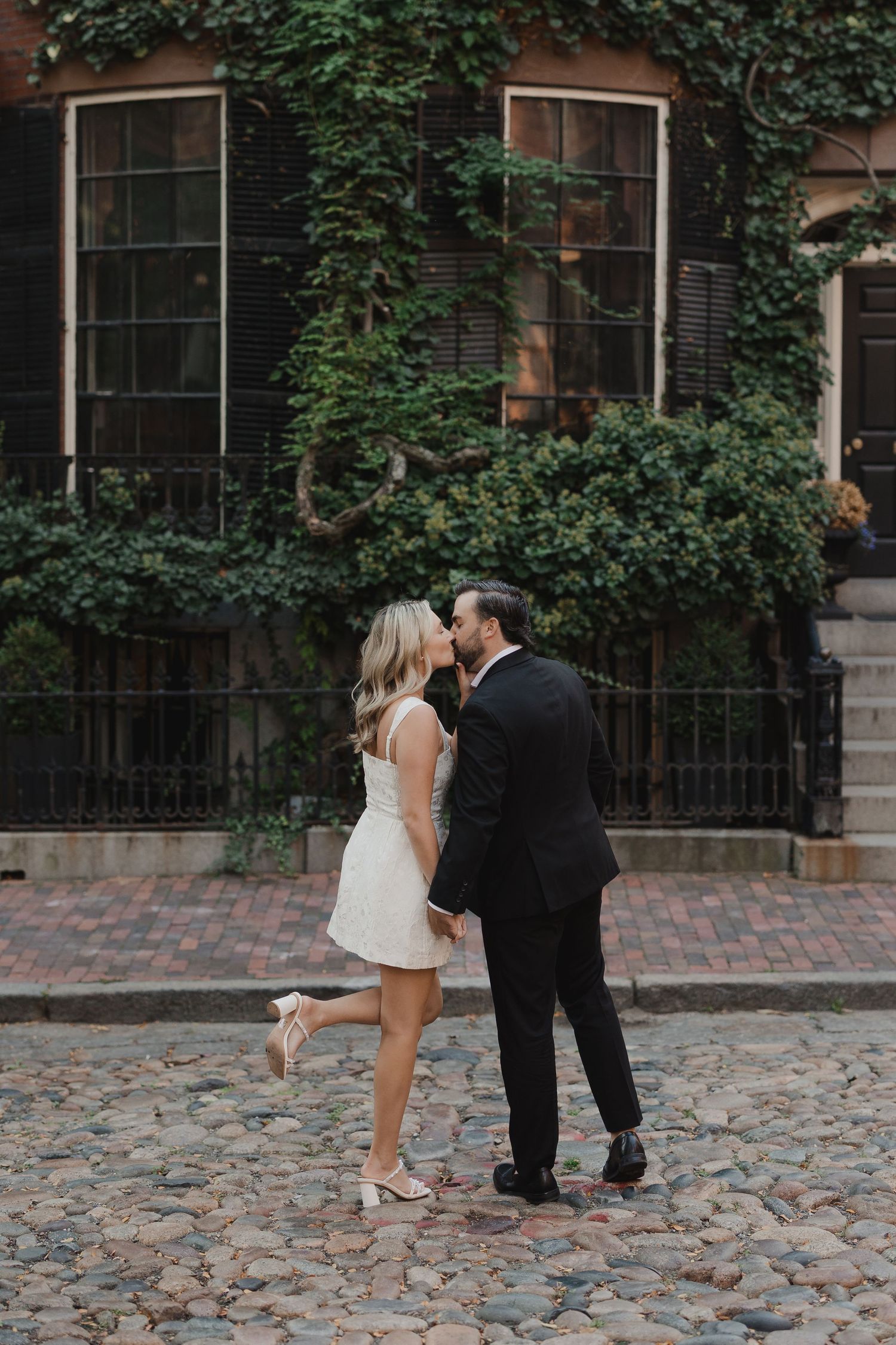 Couple sharing a kiss on cobblestone street, bride in white mini dress and groom in black suit, ivy-covered building backdrop.