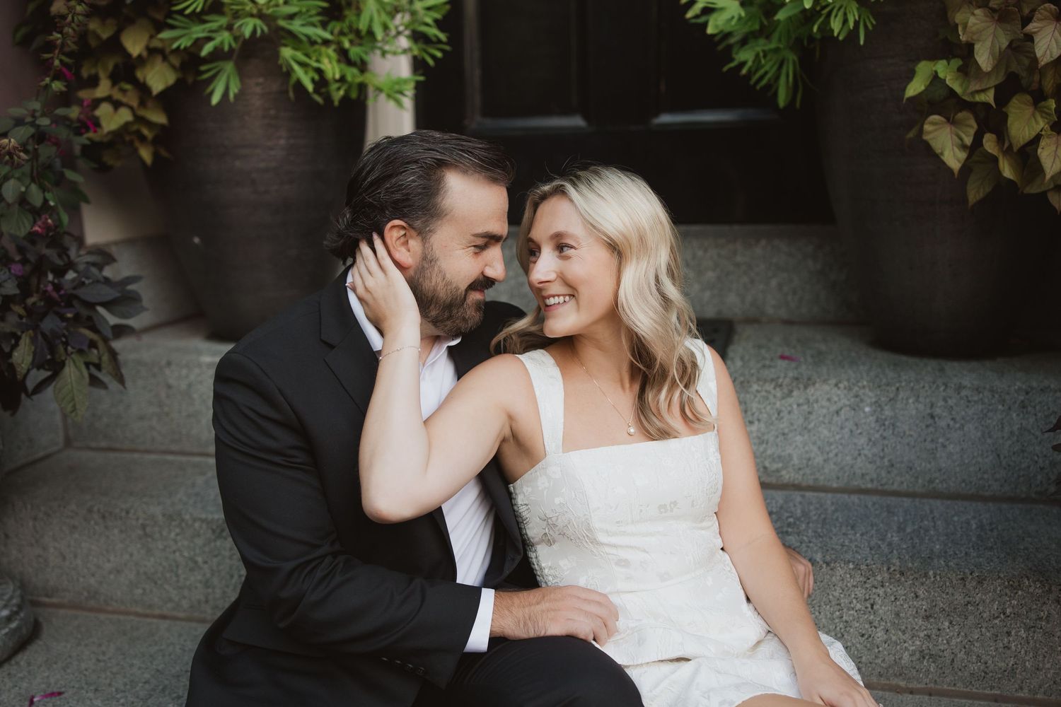 A bride and groom sit on stone steps, smiling intimately at each other in elegant wedding attire outdoors.