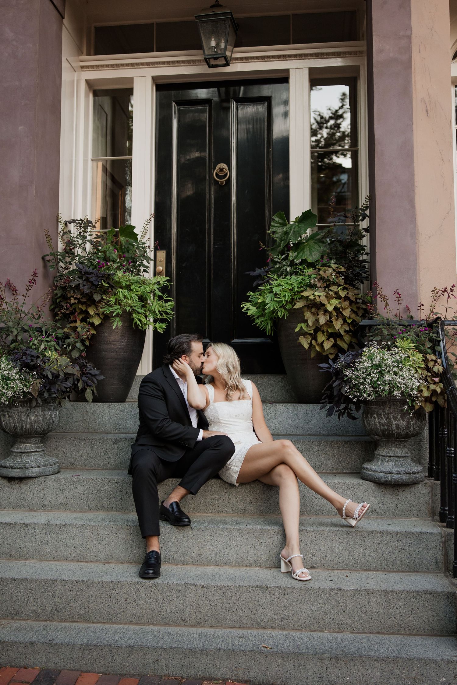 Couple kissing on brownstone steps in elegant attire, surrounded by lush potted plants and a black door.