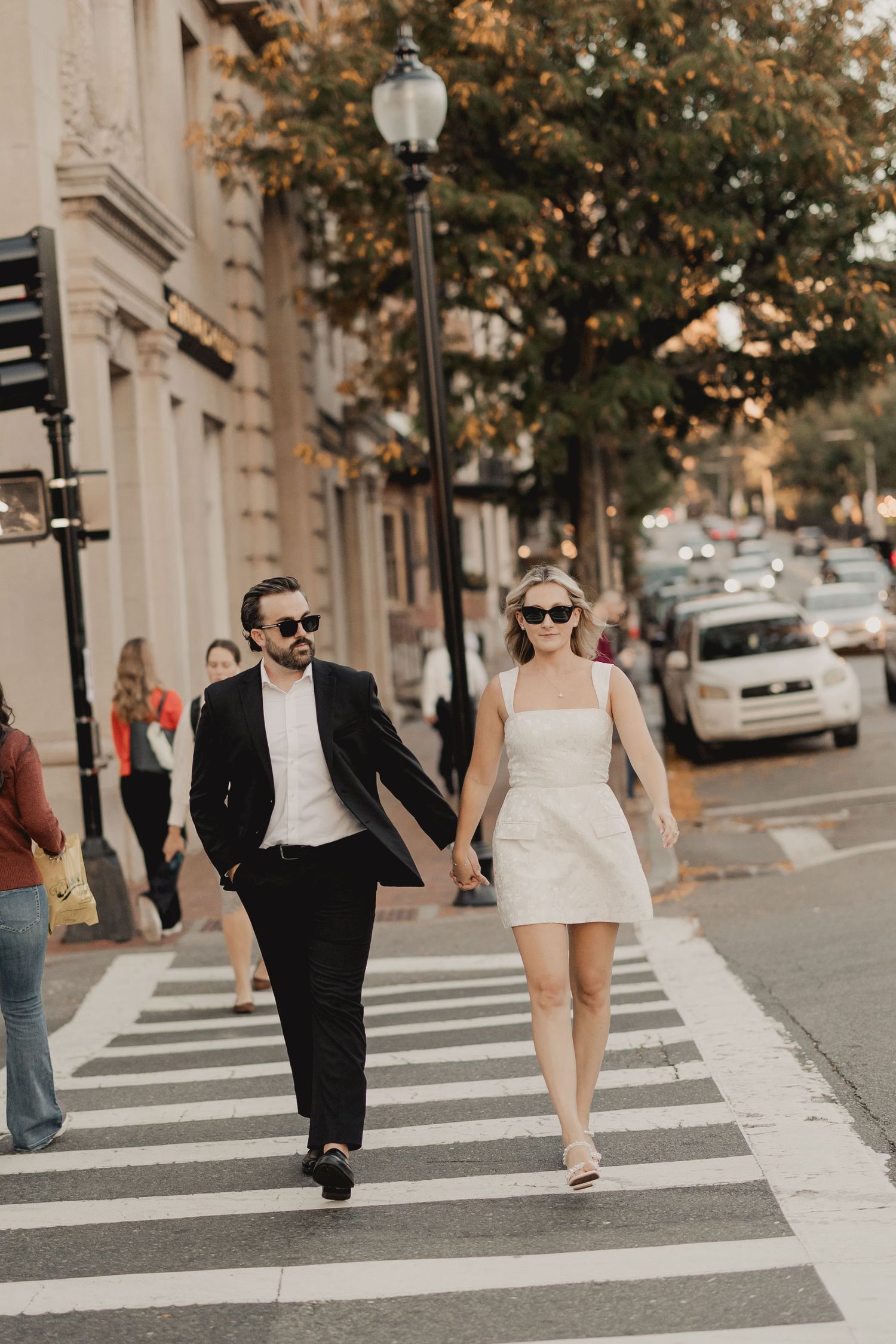 Couple in wedding attire crossing a city street crosswalk, man in black suit and woman in white mini dress.