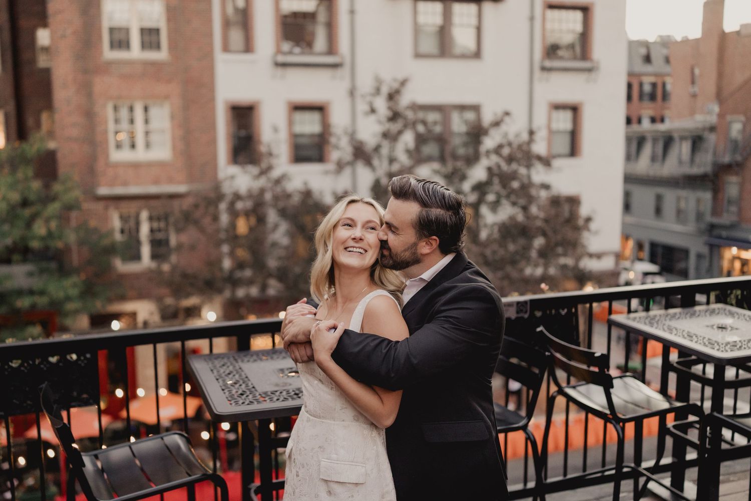 Bride and groom share romantic rooftop moment in Boston, groom kissing bride's cheek at dusk.