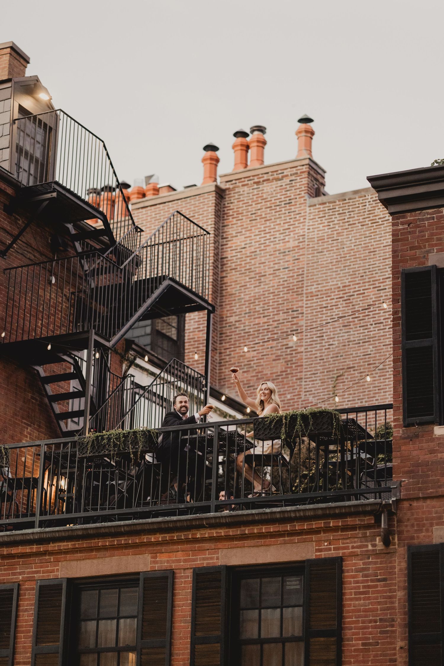 Couple laughing on a brick building balcony with fire escape stairs and chimneys in the background.