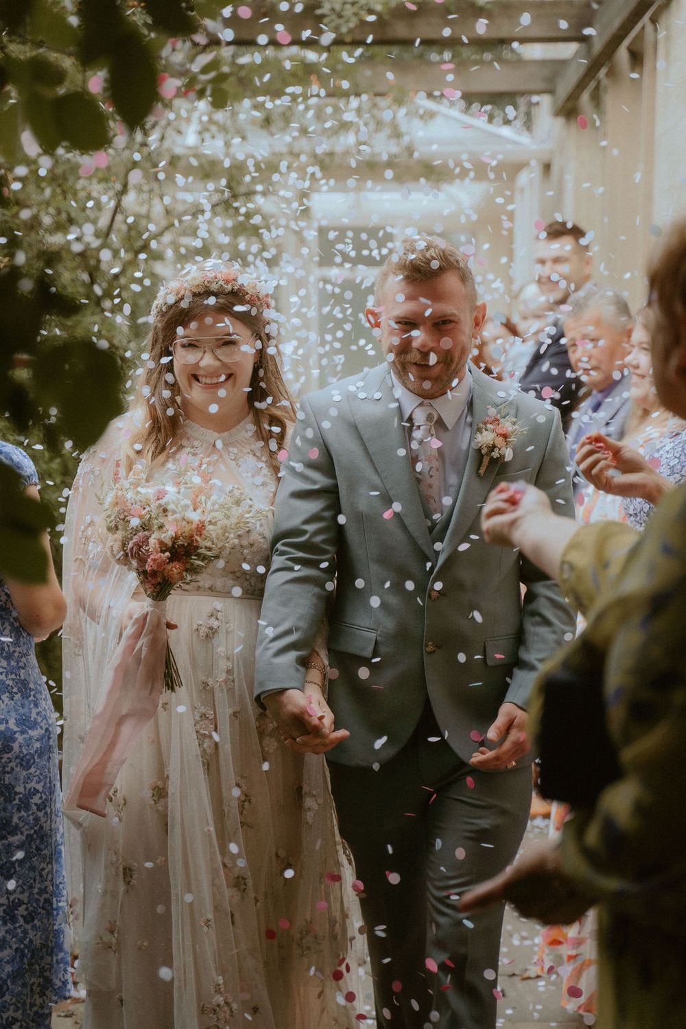 Bride in floral gown and groom in gray suit walking hand-in-hand through shower of confetti with joyful guests at outdoor ceremony