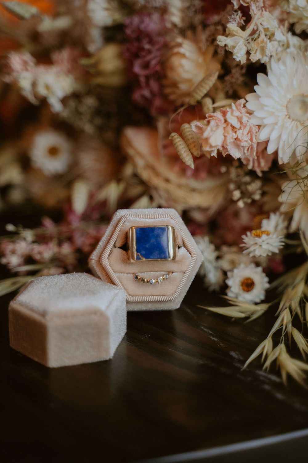 Gold ring with blue gemstone in beige hexagonal box surrounded by dried flowers in muted pink, white, and burgundy tones