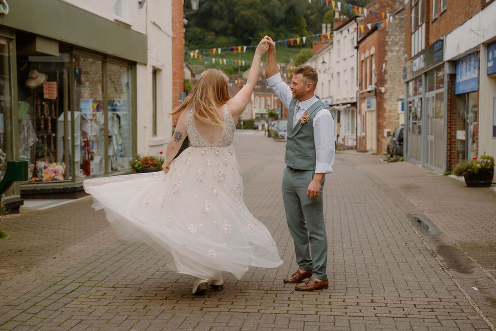 Bride in white dress twirling with groom insuit on cobblestone streets of Dursley surrounded by quaint storefronts in small town.
