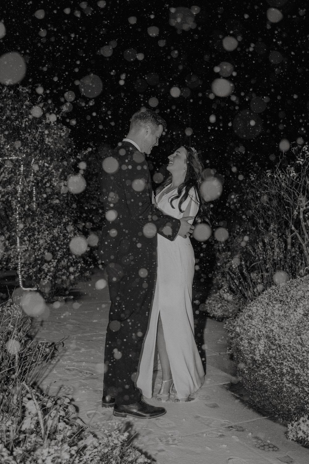 Bride and groom embracing in snowy night garden, black and white photograph with snowflakes surrounding the couple's intimate moment.