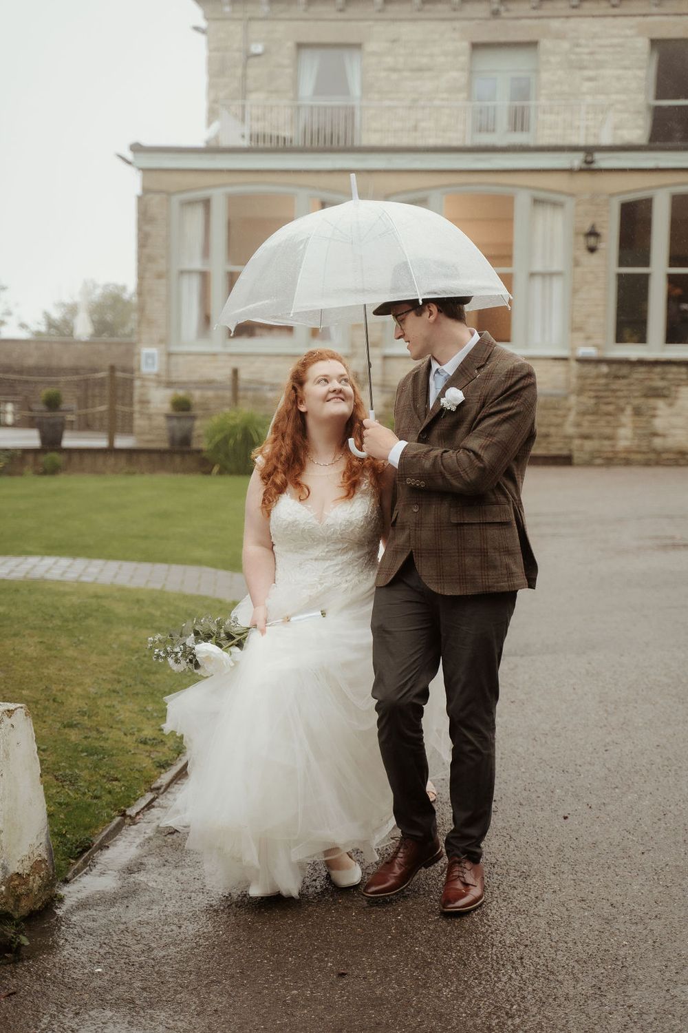 ride with flowing gown and groom in brown suit sharing white umbrella outside stone venue, bride holding bouquet on rainy day.