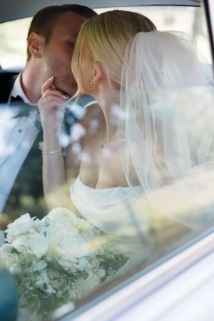 Bride and groom share an intimate kiss in a car as her white veil flows and bouquet rests nearby.