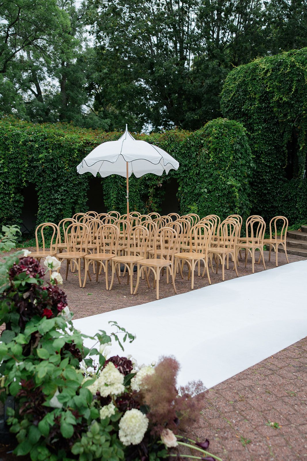 Outdoor wedding ceremony setup with rattan chairs, white aisle runner, elegant parasol, and lush green hedges backdrop.