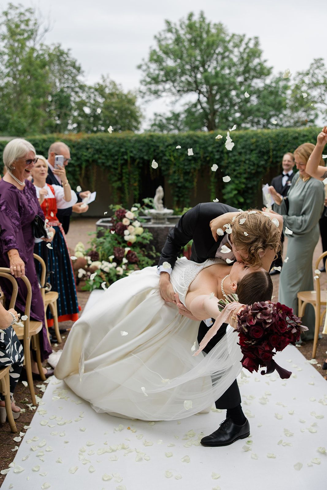 Groom dips bride for a kiss on white aisle runner at outdoor wedding ceremony with guests cheering around them.