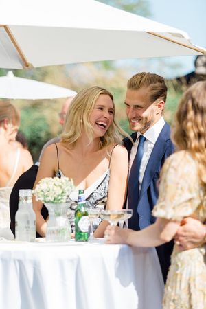 A group of wedding guests share joyful laughter while seated at an outdoor reception table under blue skies.