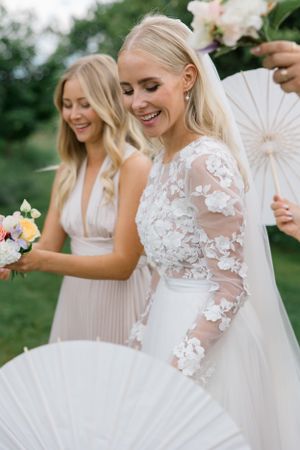 Happy wedding party members with delicate white fans and bouquets celebrate outdoors.
