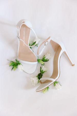 Elegant white bridal sandals with strappy design decorated with small green and white floral accents on a light background.