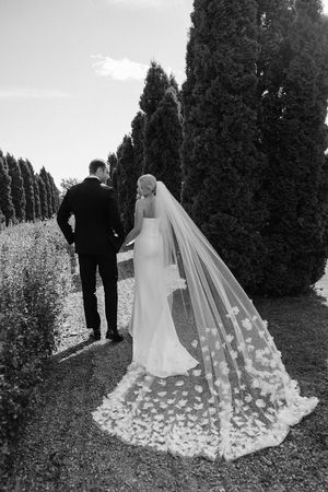 A bride and groom walk along a cypress tree-lined path, with her long floral-detailed veil trailing behind them.