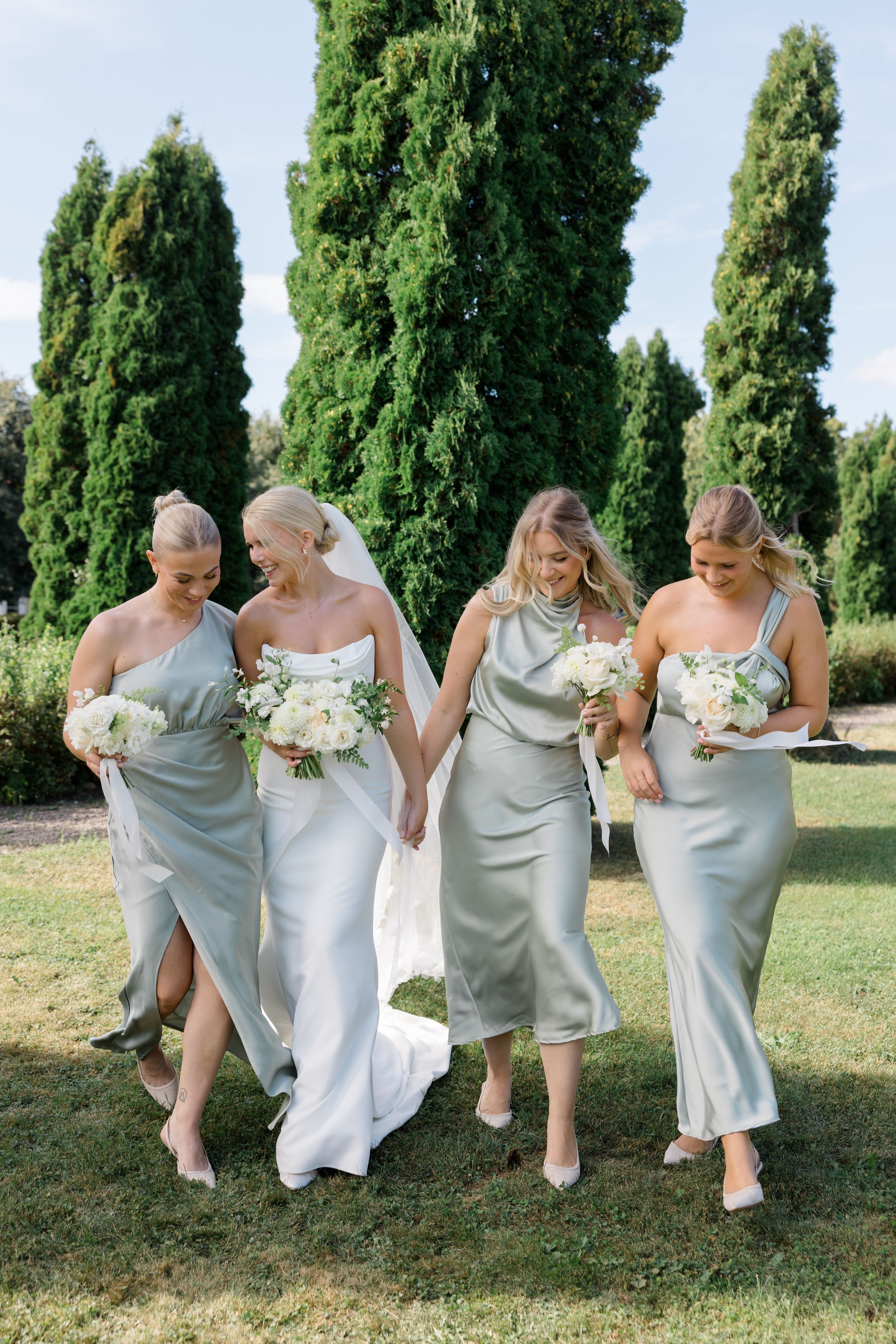 A bridal party in silver and white dresses walks together on grass with tall cypress trees in the background.