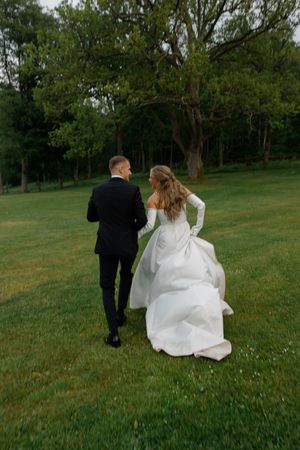 A couple in formal wedding attire walks together across a lush green lawn surrounded by trees in the background.