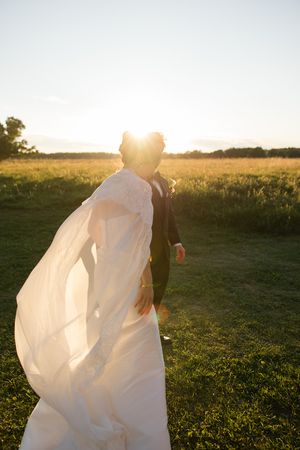 A wedding couple stands in a golden field at sunset as the bride's white dress and veil flow in the breeze.