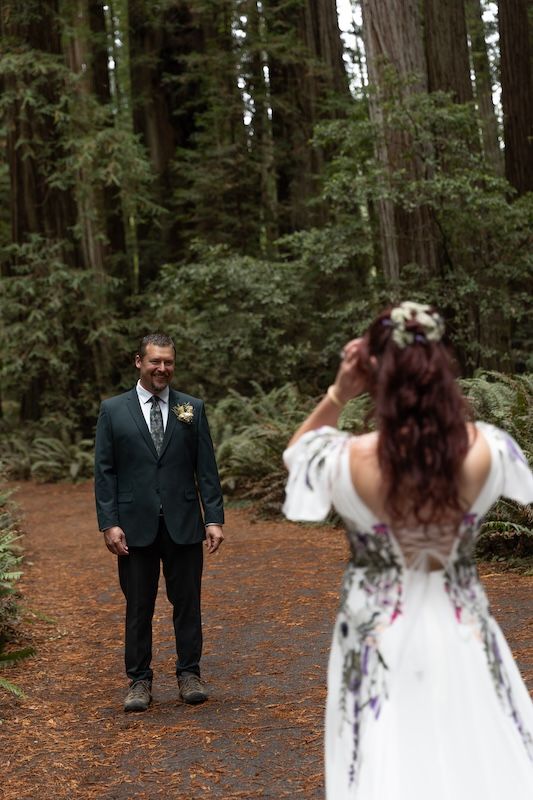 Bride in floral wedding dress faces groom in green suit during first look ceremony in lush redwood forest.