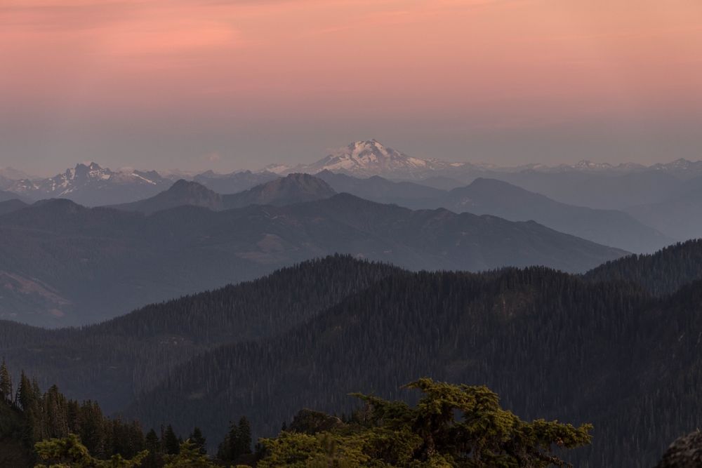 Stunning sunset over layered mountain ridges with snow-capped peaks glowing pink in the twilight sky.