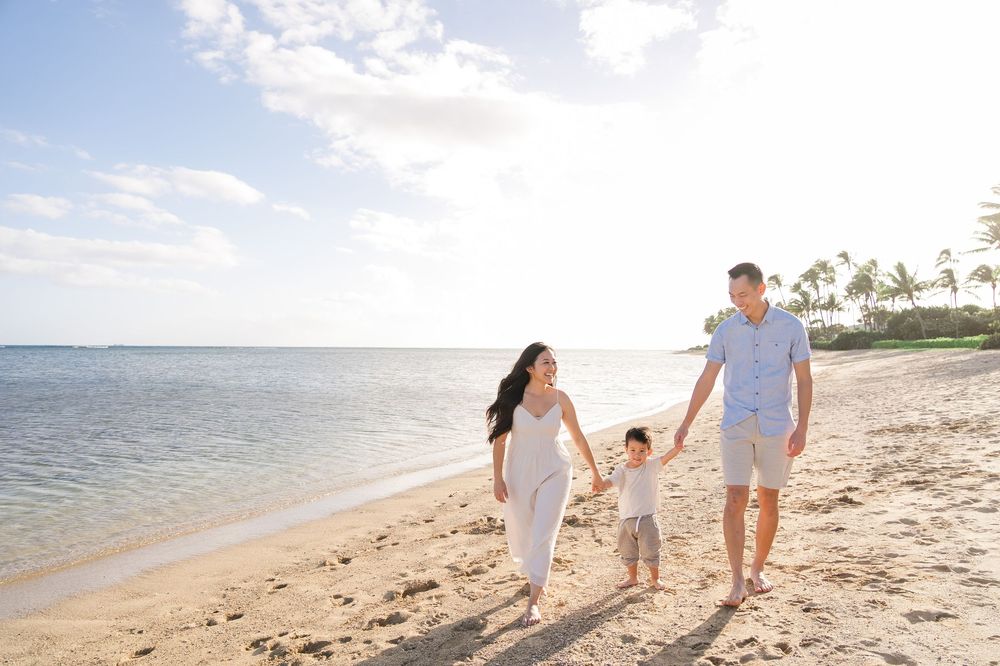 A family walks hand in hand along a serene beach at sunset with palm trees in the distance.