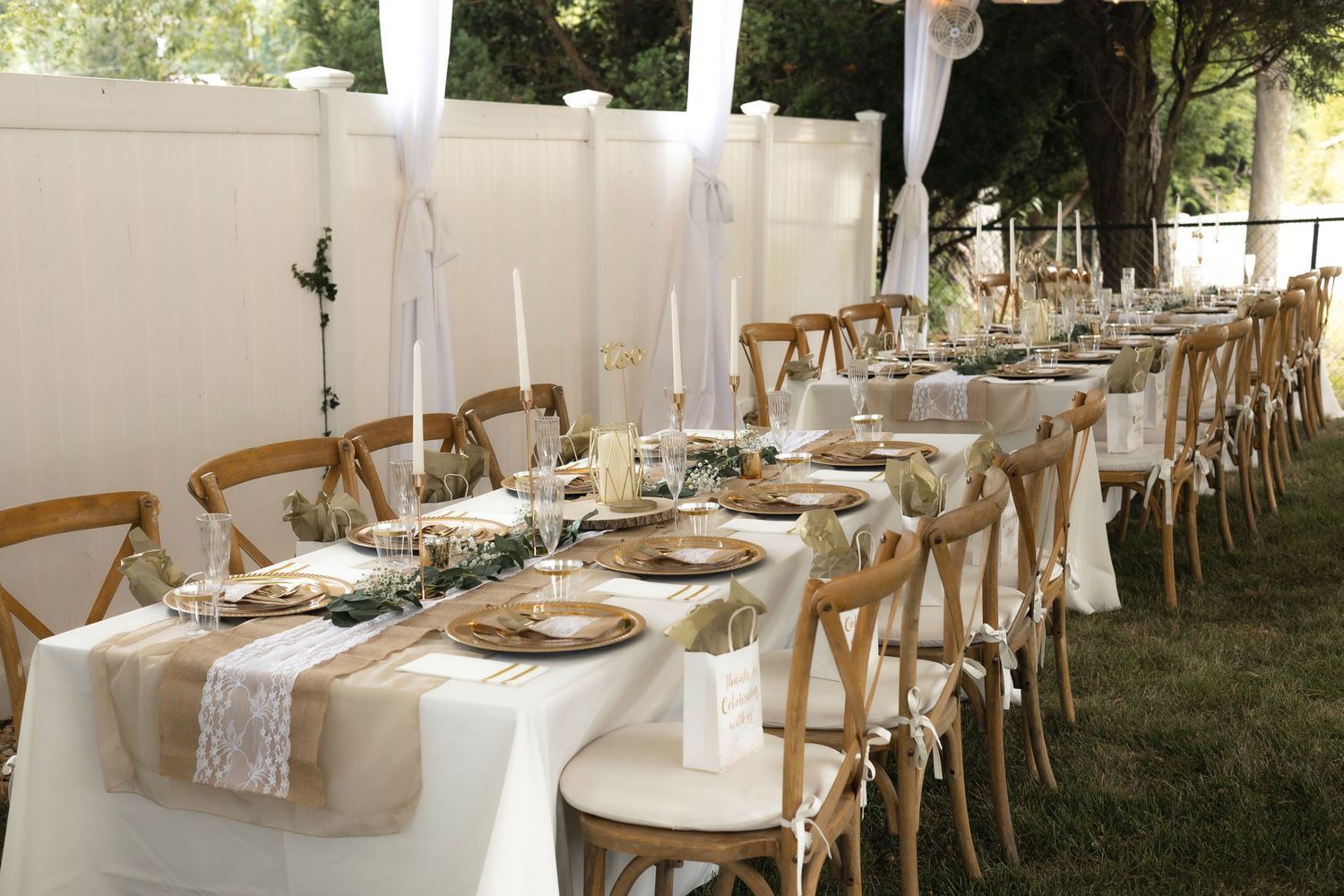 Elegant outdoor wedding table setting with white linens and wooden chairs under sheer drapes.