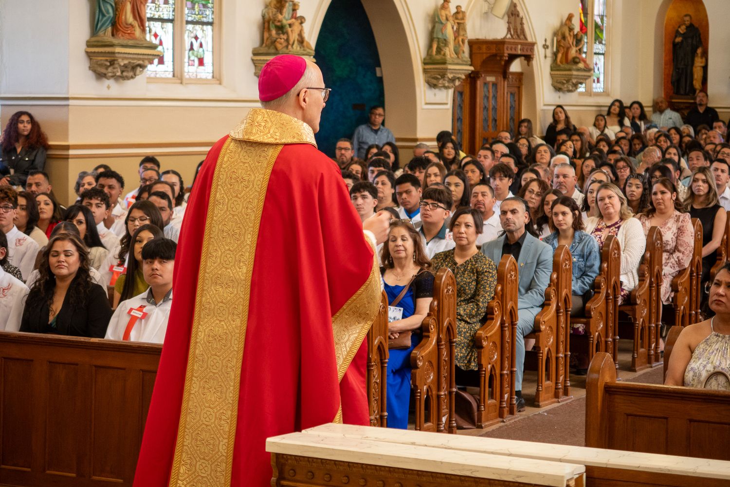 Clergy member in red and gold vestments addresses a full congregation inside an ornate church sanctuary during service.