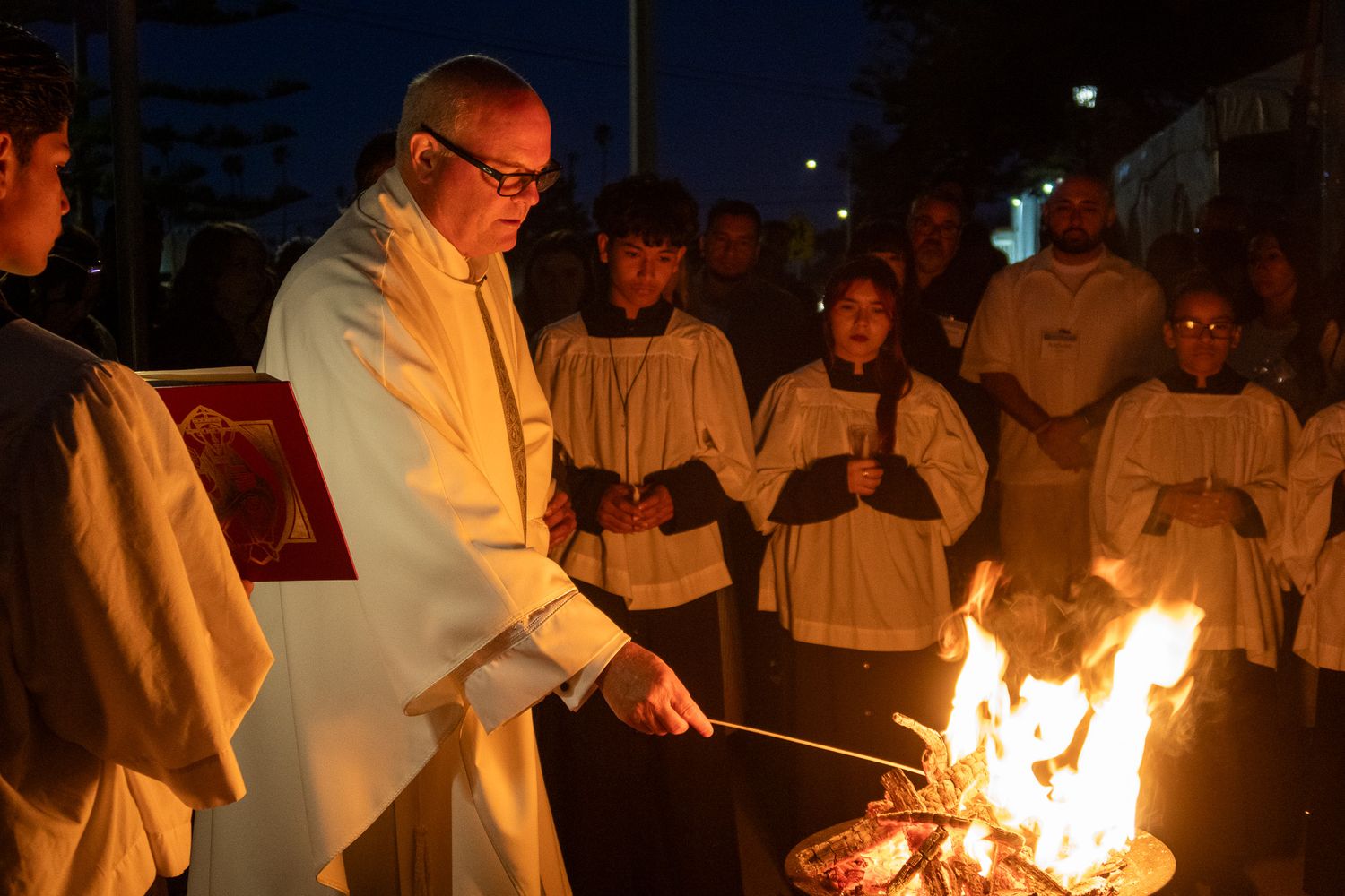 A group gathers around a ceremonial fire at night with a person in white robes lighting it.