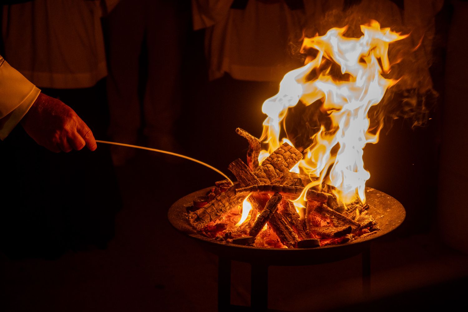 A hand holds a stick near bright flames burning in a metal fire pit during a nighttime ceremony.