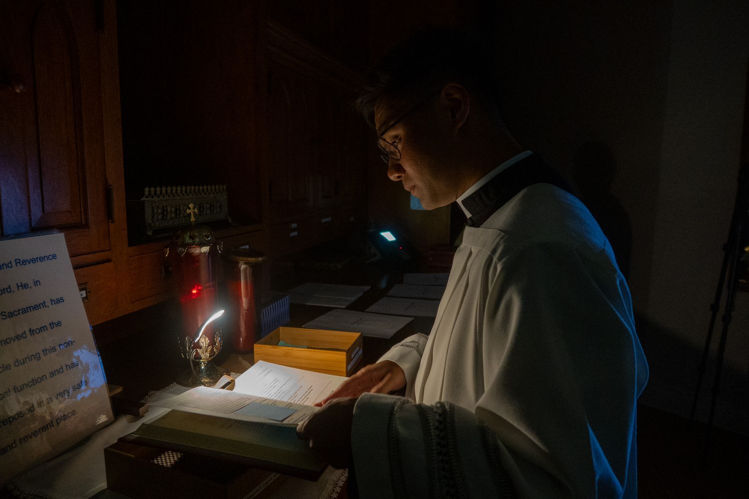 Someone reads by candlelight in dark robes against a dimly lit wooden interior.