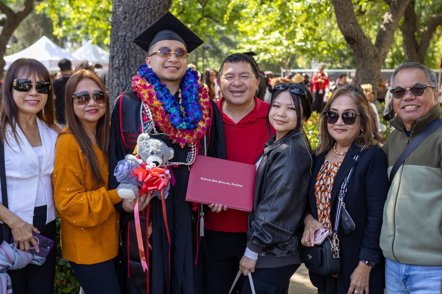 A group of people smiling outdoors at a graduation celebration with trees in the background.