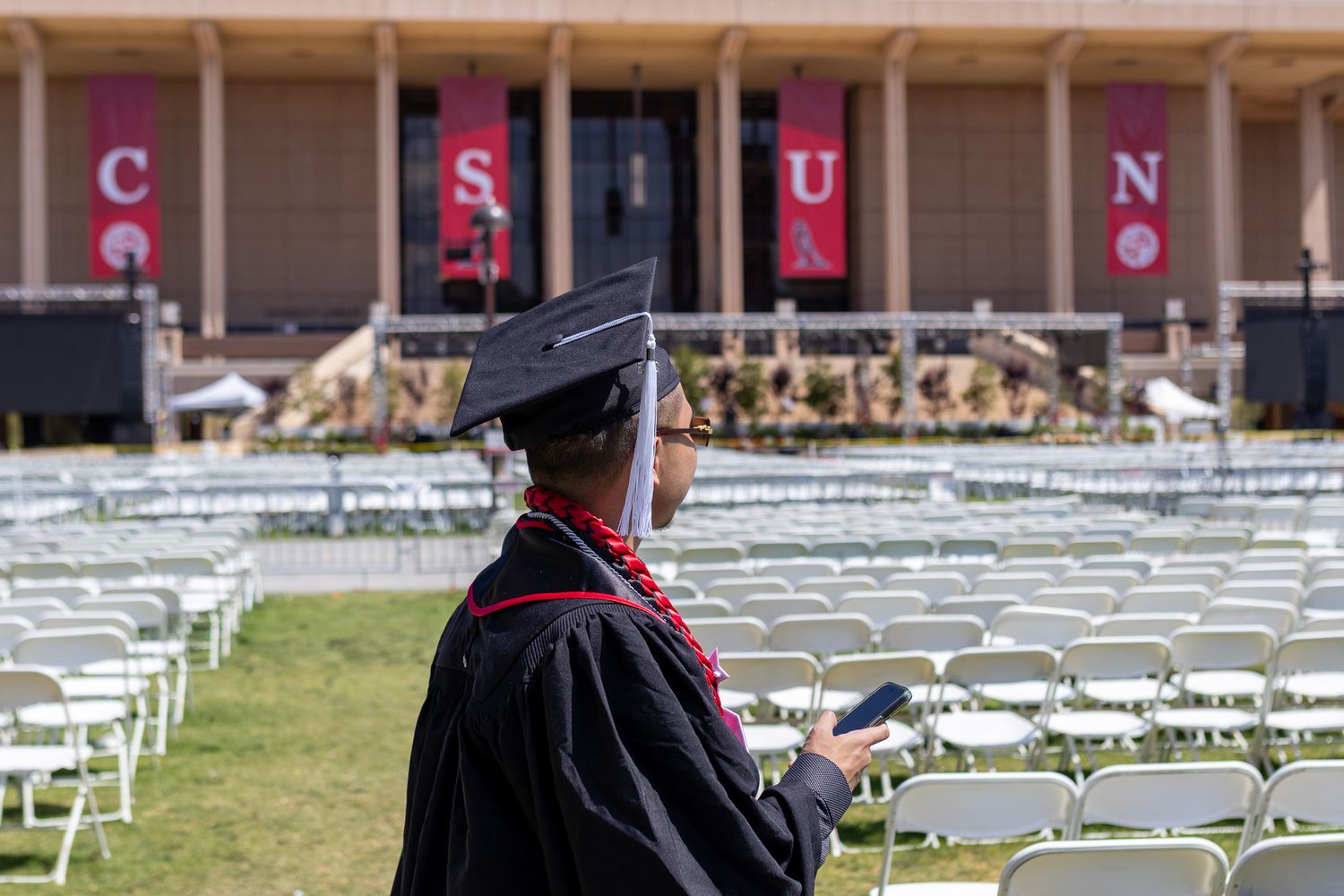 Graduate in cap and gown stands before rows of white chairs at outdoor commencement ceremony.