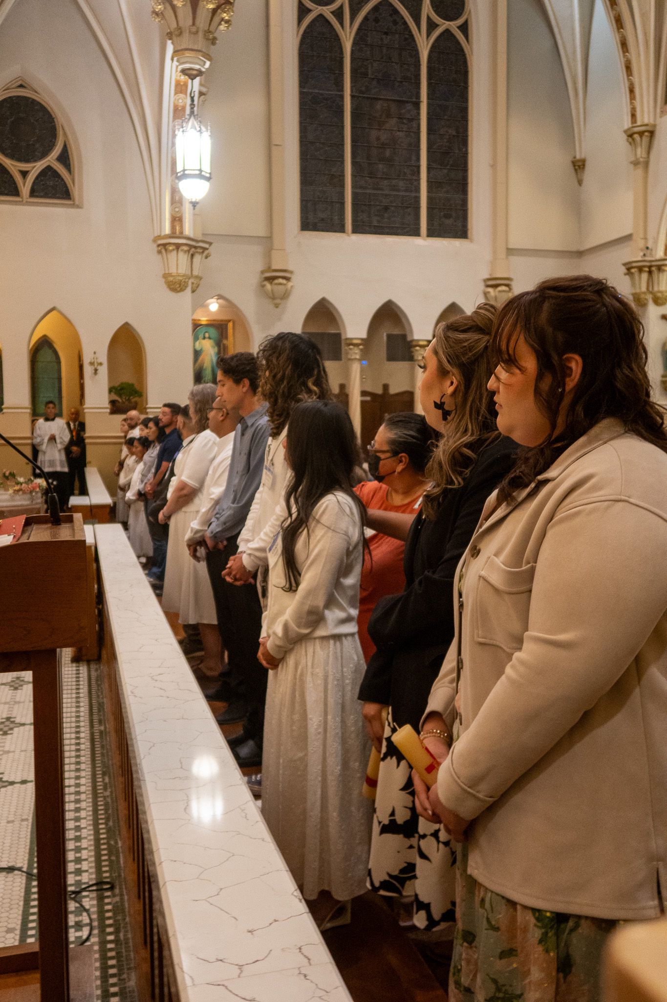 People stand in a row during a church service inside a Gothic-style sanctuary with arched windows and hanging lanterns.