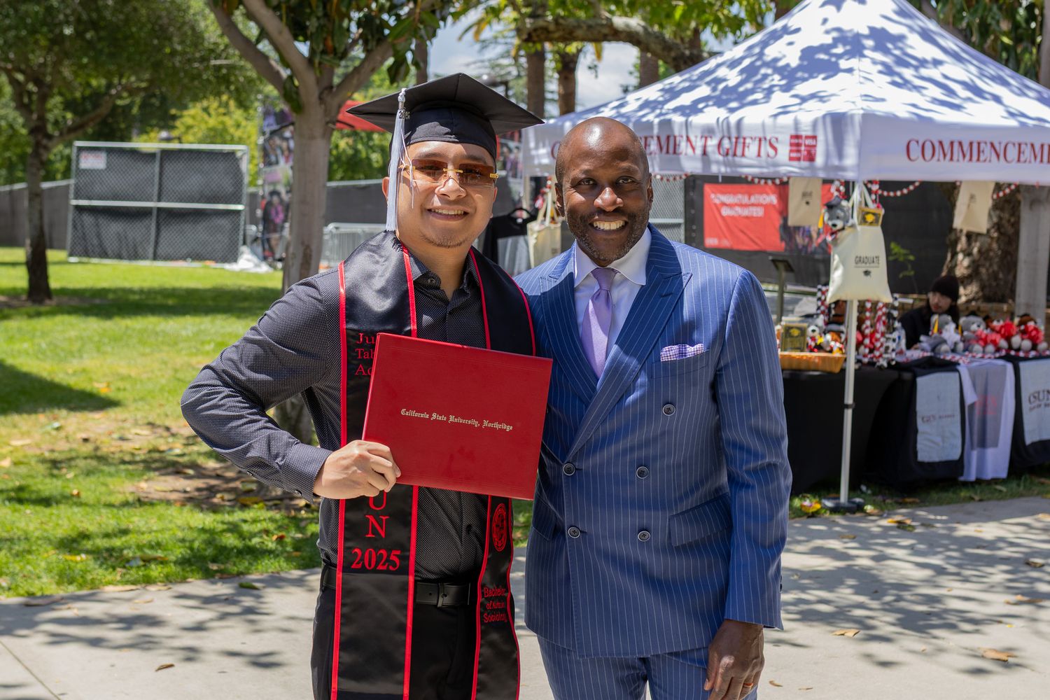 Two people in formal attire pose together at an outdoor graduation celebration with tents in the background.