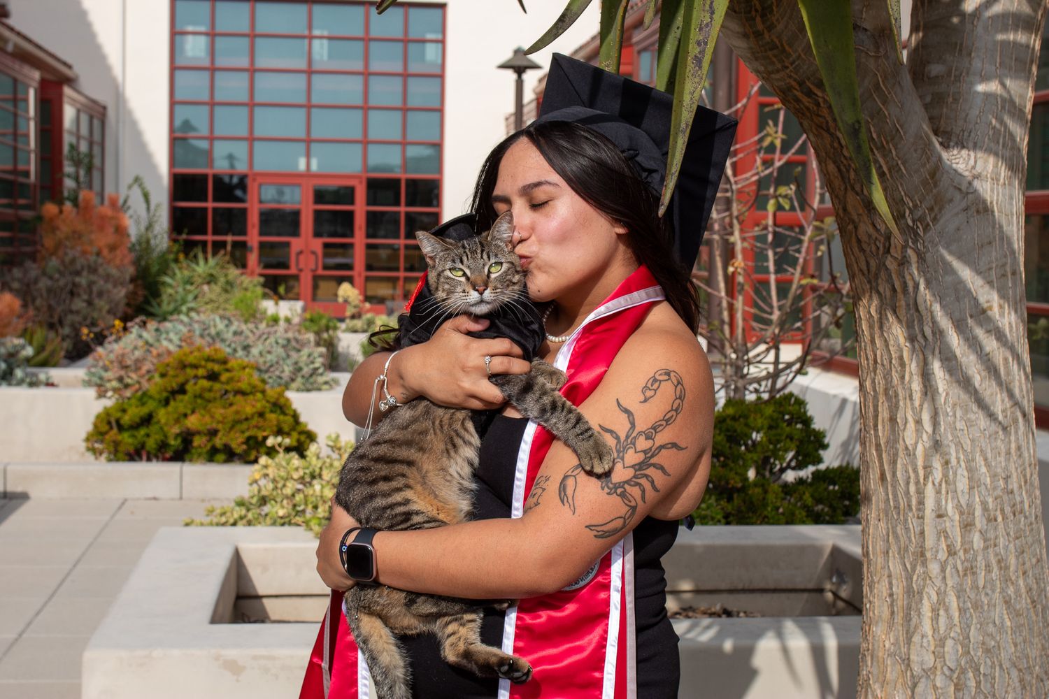 A person in red and black clothing holds and cuddles a grey striped cat outdoors near a building.