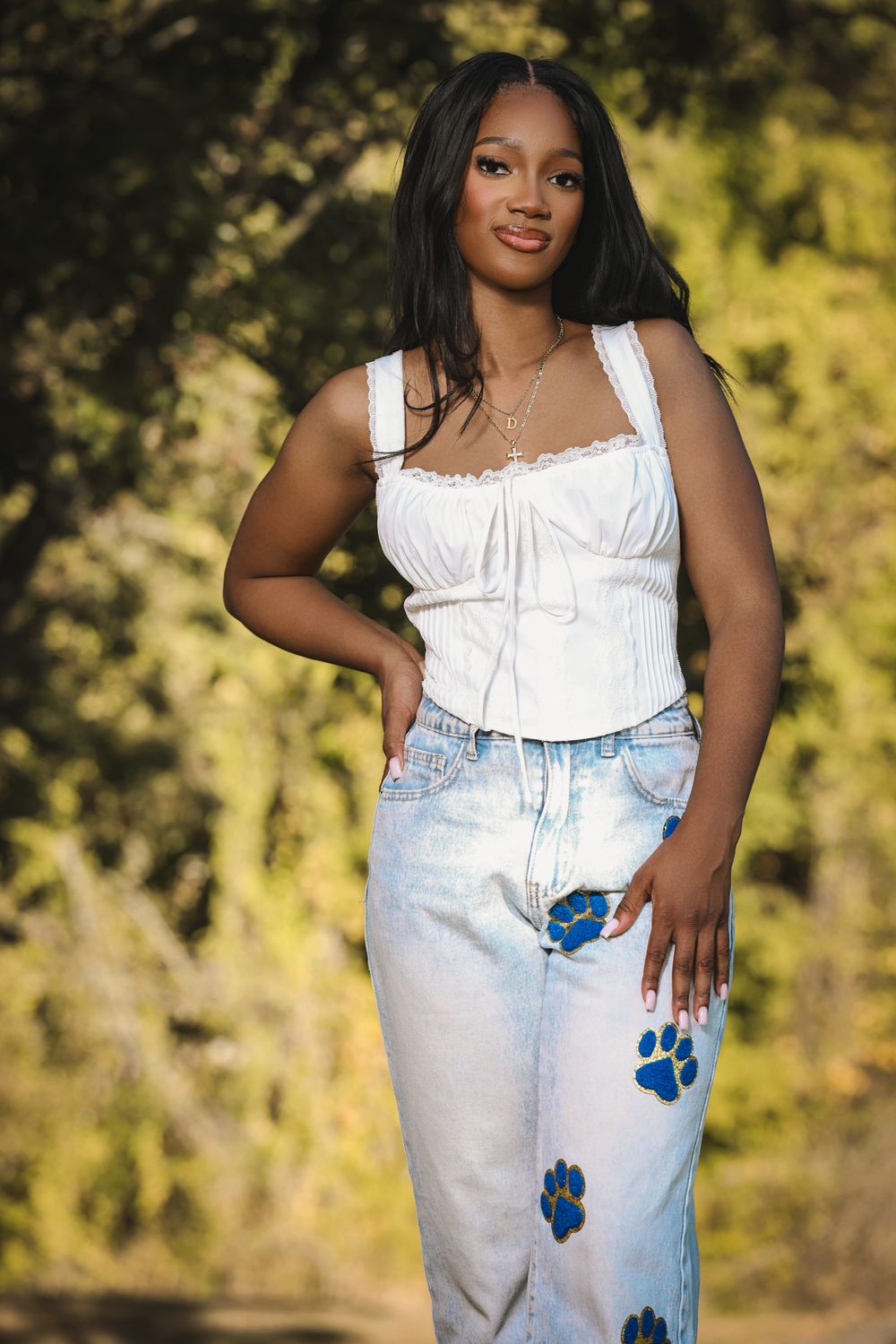 Young woman in white corset top and blue paw print jeans posing outdoors with green trees in background.