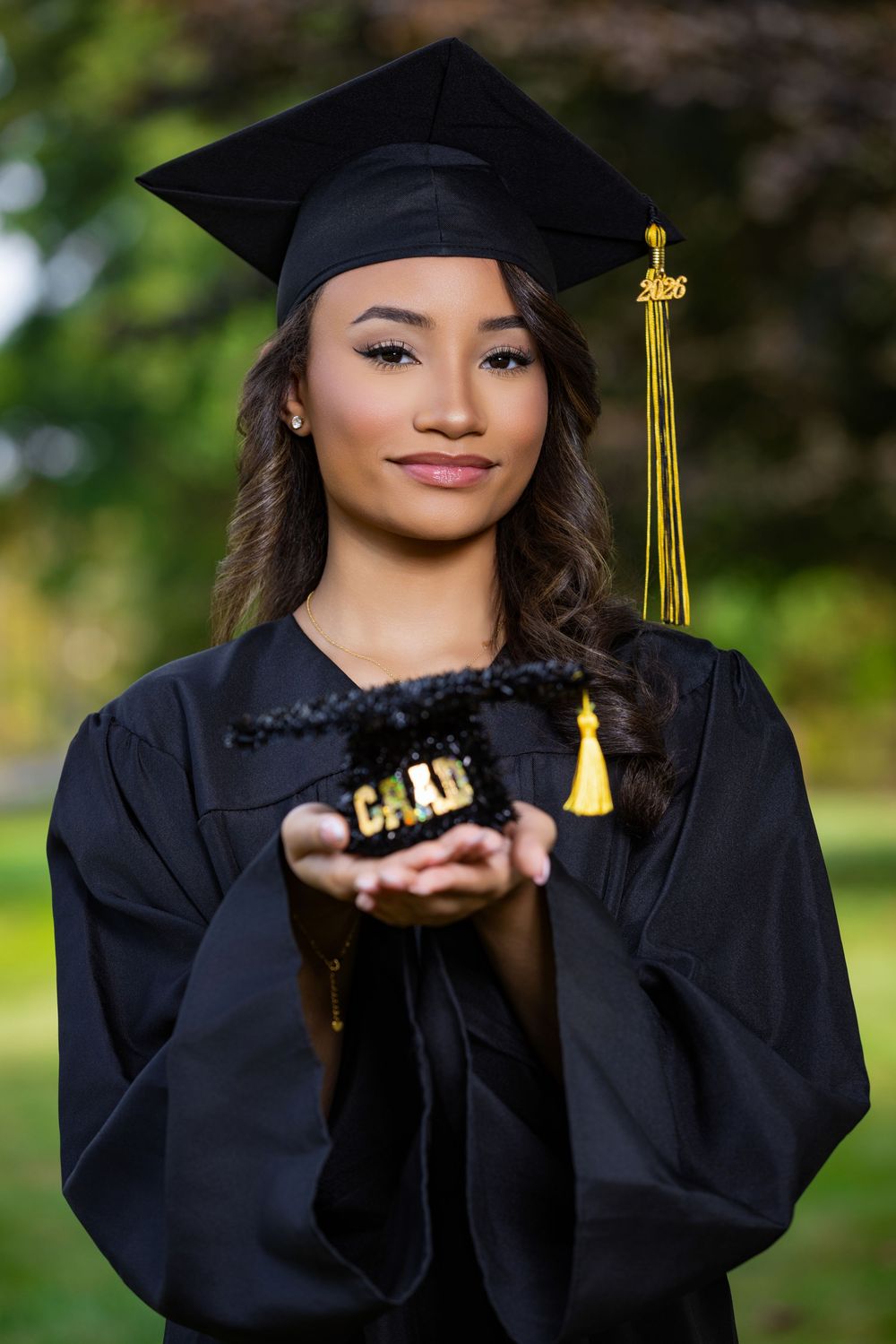 Young woman in black graduation cap and gown holds mini grad cap, smiling outdoors.