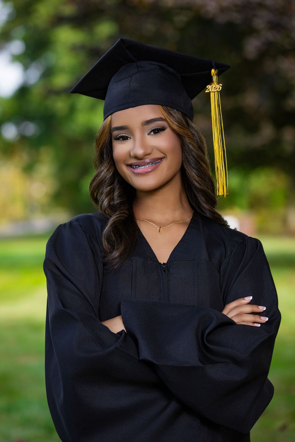 Smiling young woman in black graduation cap and gown with gold tassel, arms crossed, posing outdoors.