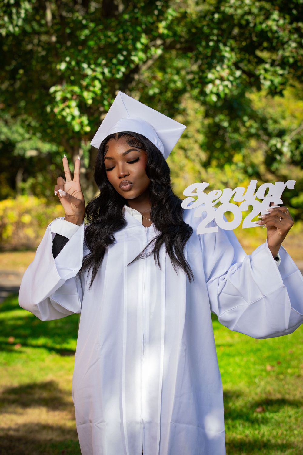 Young woman in white cap and gown holds Senior 2024 sign while flashing peace sign outdoors.