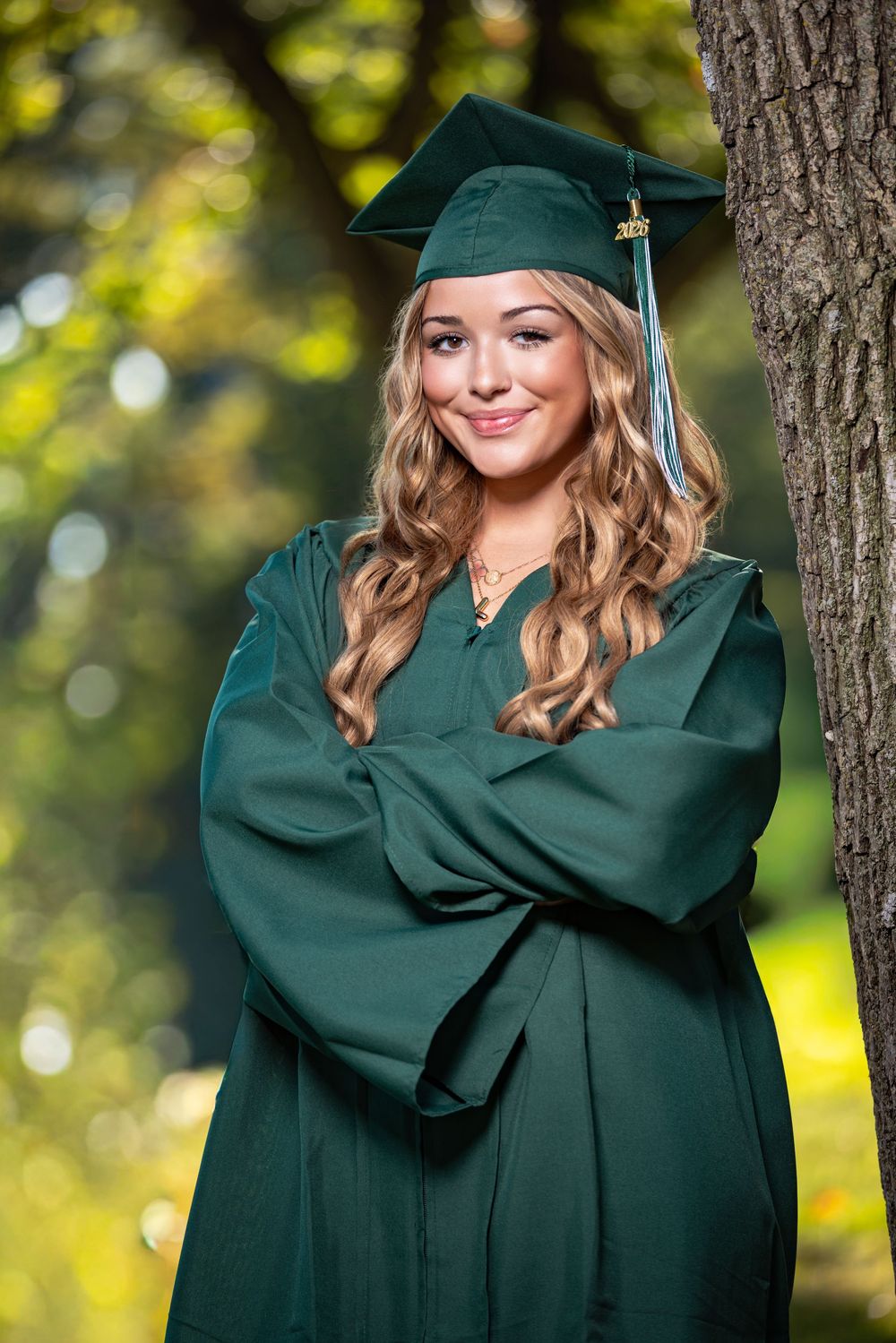 Smiling young woman in green graduation cap and gown leaning against a tree in a sunlit outdoor setting.