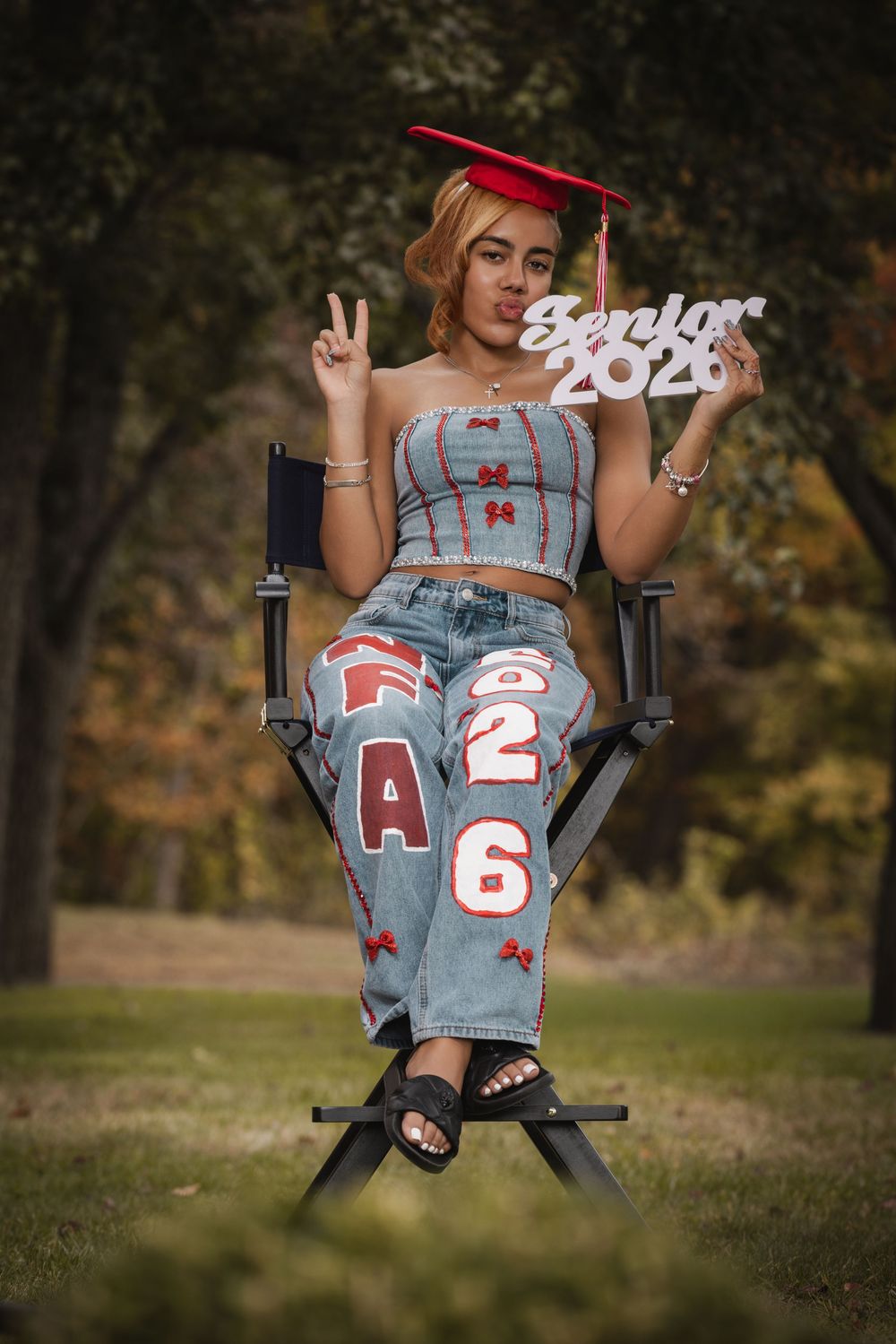 A young woman in a red graduation cap holds a Senior 2026 sign while seated outdoors in a director's chair.