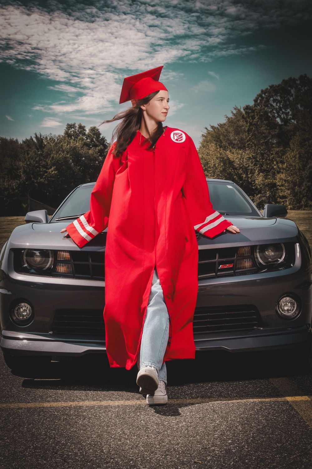 A young woman in a red graduation cap and gown poses proudly in front of a gray Chevrolet Camaro outdoors.