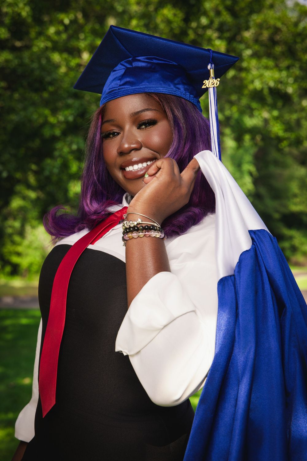 Smiling graduate in blue cap and gown with purple hair posing outdoors among green trees.