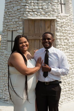 A couple dressed in formal attire pose and smile together against a white stone wall with natural lighting.